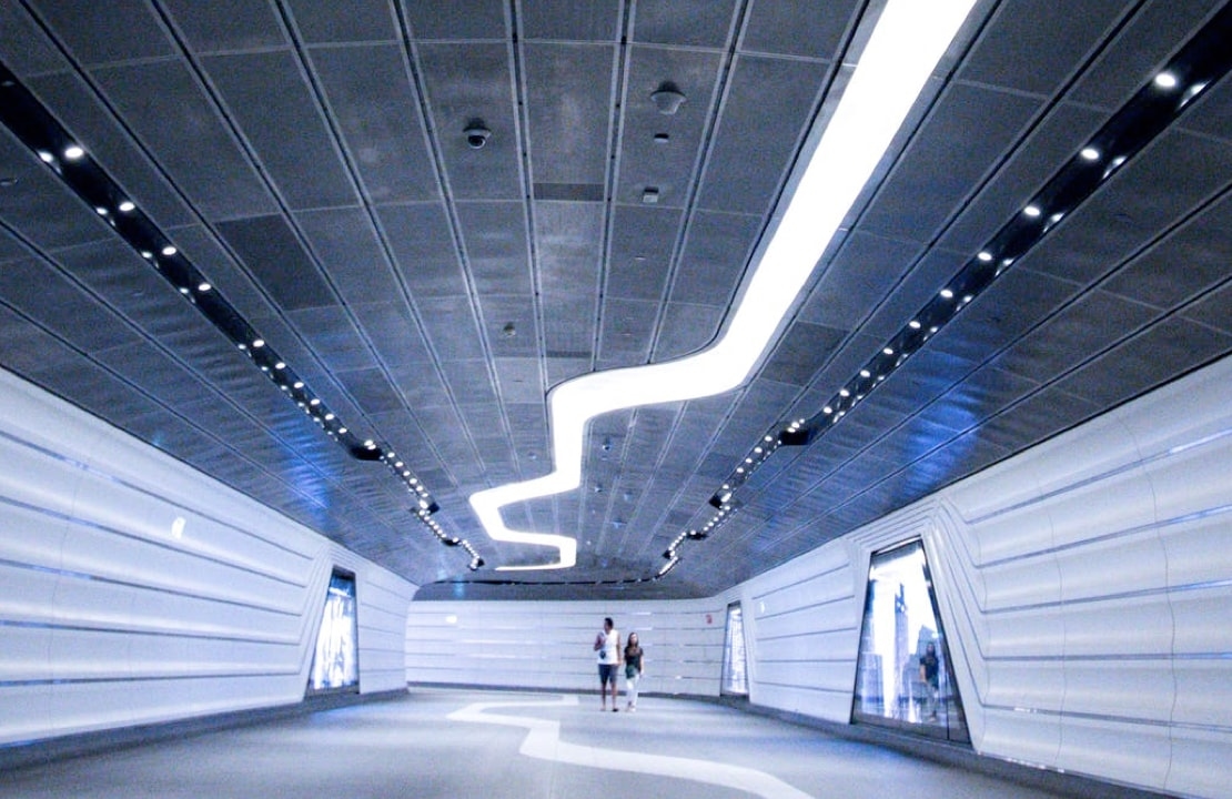 A futuristic-looking tunnel with white walls, a curved ceiling, and bright, linear lights illuminating the path.