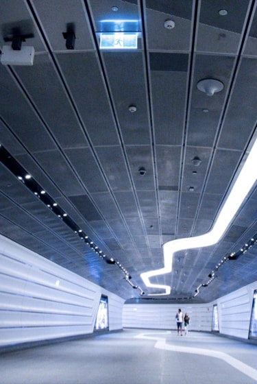 A futuristic-looking tunnel with white walls, a curved ceiling, and bright, linear lights illuminating the path.