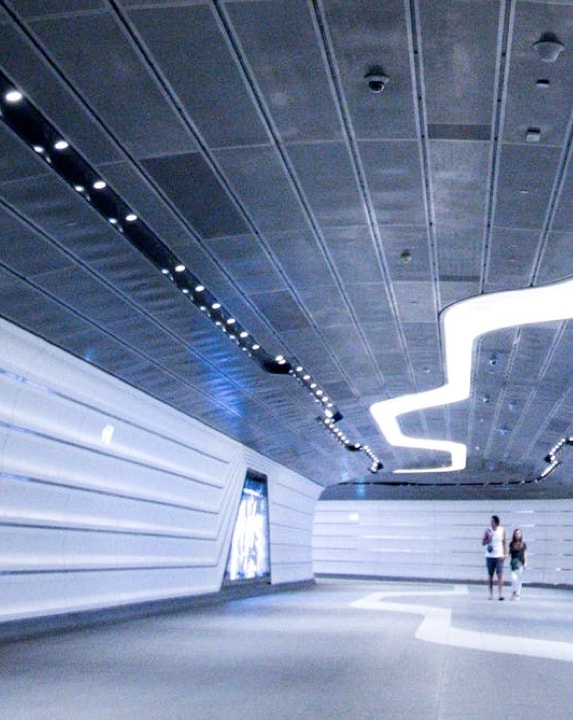 A futuristic-looking tunnel with white walls, a curved ceiling, and bright, linear lights illuminating the path.