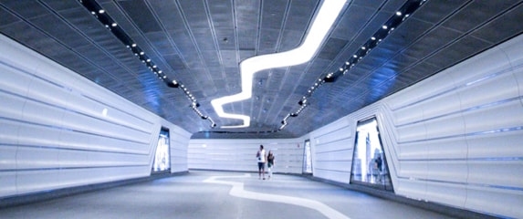 A futuristic-looking tunnel with white walls, a curved ceiling, and bright, linear lights illuminating the path.