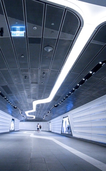 A futuristic-looking tunnel with white walls, a curved ceiling, and bright, linear lights illuminating the path.