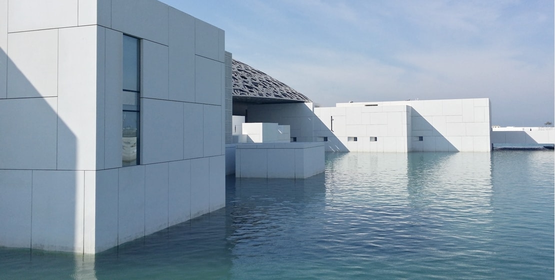 A low-angle view of a white, modern building with a geometric, lattice-like roof. The building is partially submerged in a body of water, creating a unique architectural design.