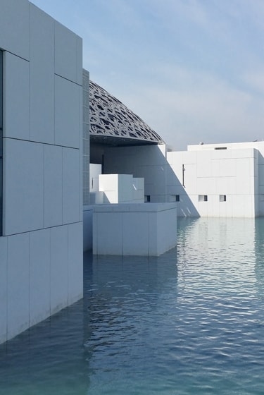 A low-angle view of a white, modern building with a geometric, lattice-like roof. The building is partially submerged in a body of water, creating a unique architectural design.